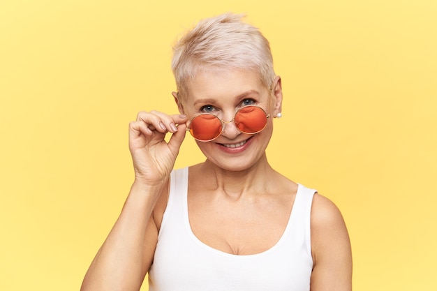 Studio shot van trendy glamoureuze vrouw van middelbare leeftijd met pixie kapsel poseren geïsoleerd haar stijlvolle ronde zonnebril opstijgen.