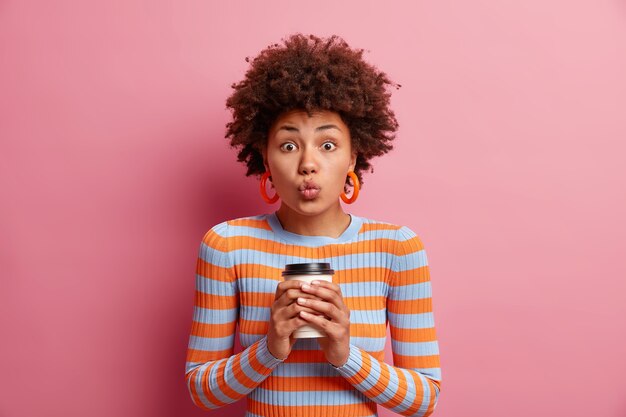 Studio shot van verrast mooie jonge vrouw houdt lippen gevouwen drankjes afhaalmaaltijden koffie wil je kussen draagt oranje oorbellen gestreepte trui geïsoleerd over roze muur.