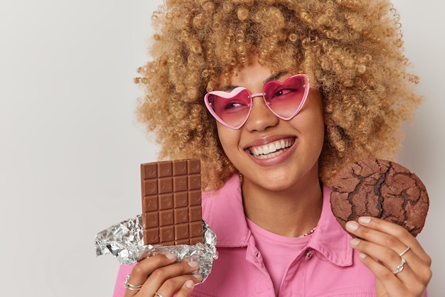 Gratis foto studio shot van vrolijke jonge vrouw heeft zoetekauw houdt reep chocolade en cookie kiest tussen twee desserts glimlacht breed draagt roze zonnebril en jas geïsoleerd op witte achtergrond