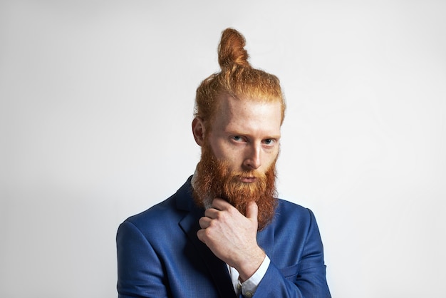 Studio shot van zelfverzekerde stijlvolle jonge roodharige zakenman met haarknotje en wazige baard, hand op kin houden en camera kijken met peinzende uitdrukking, na te denken over zakelijke ideeën en plannen