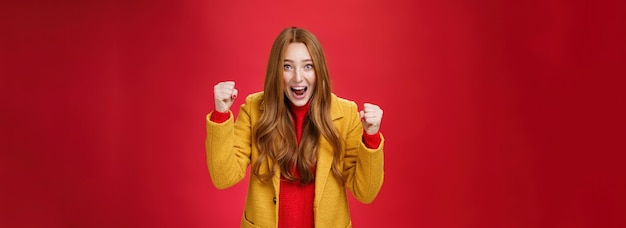 Gratis foto studio shto van een vrolijk opgewonden knap rood meisje in een gele jas die gebalde vuisten opheft van vreugde