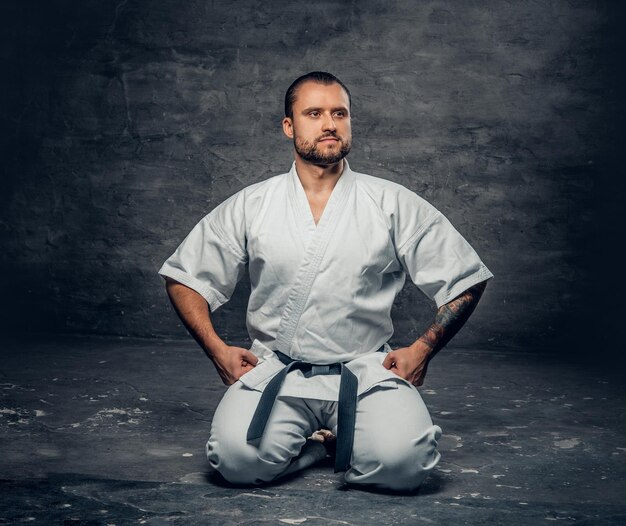 Studioportret van de bebaarde brutale karatevechter gekleed in een witte kimono over grijze achtergrond.