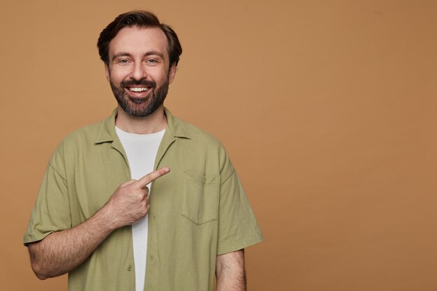 Gratis foto studioportret van een bebaarde man die zich voordeed op beige achtergrondpunten met een vinger opzij op kopieerruimte met een brede glimlach op zijn gezicht