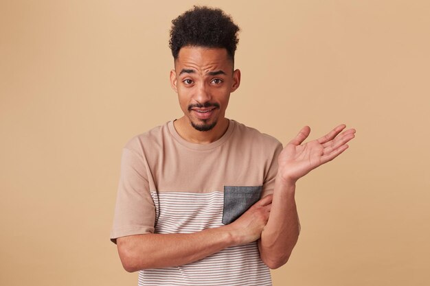 Studioportret van een jonge afro-amerikaanse man die rechtstreeks in de camera kijkt met een geïrriteerde gezichtsuitdrukking, zijn hand opsteekt, poserend over een beige achtergrond