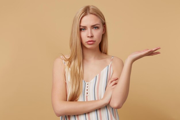 Studioportret van een jonge blonde vrouw die zich voordeed op een beige achtergrond, stak haar hand op met een geïrriteerde gezichtsuitdrukking