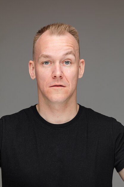 Studioportret van een knappe man met kort blond haar en blauwe ogen in een zwart t-shirt die naar de camera kijkt met een wenkbrauw omhoog, zelfvertrouwen en arrogantie toont.