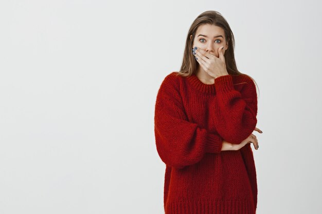 Studioportret van geschokte verbaasde aantrekkelijke vrouw in losse sweater, die geopende mond met palm bedekt en doodsbang staart, verbaasd en verrast