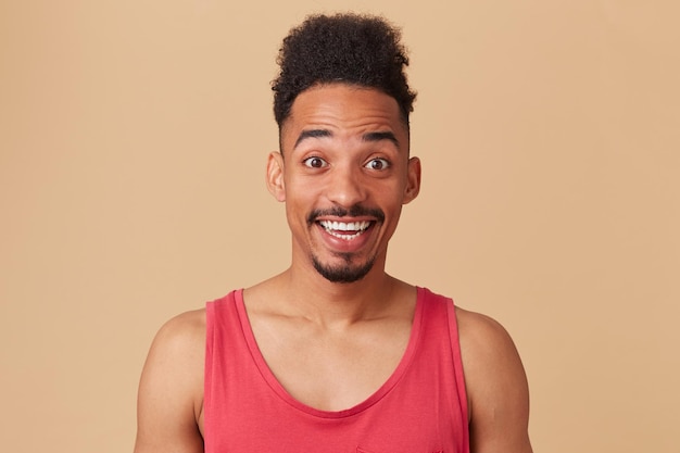 Studioportret van jonge afro-amerikaanse man houdt zijn ogen gesloten en glimlacht, lacht breed met positieve gezichtsuitdrukking die zich voordeed op beige achtergrond