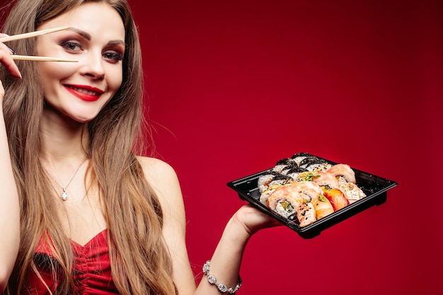 Studioportret van positief glimlachend donkerbruin meisje in rood met rode lippen en oogmake-up die heerlijke reeks Japanse sushi en stokken houdt Heldere rode achtergrondruimte voor tekst