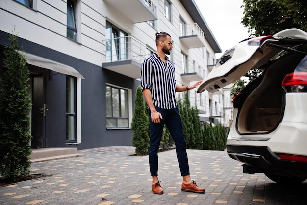 Succesvolle arabische man draagt een gestreept shirt en een zonnebril gesloten kofferbak van zijn witte suv-auto Stijlvolle arabische mannen in transport