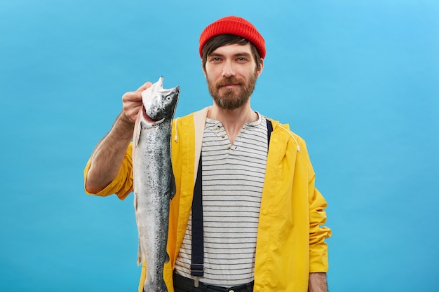 Gratis foto succesvolle bebaarde visser die zich over blauwe muur met zijn vangst bevindt die gelukkige uitdrukking heeft. knappe jongeman die lange zware vis in handen houdt die trots en opgewonden voelt