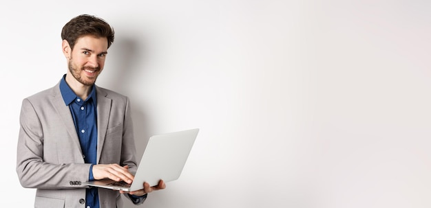 Succesvolle glimlachende zakenman die aan laptop werkt en gelukkig naar camera kijkt die zich in grijs kostuum o bevindt