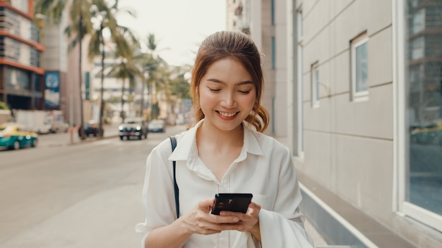 Succesvolle jonge Aziatische onderneemster in de kleren van het manierbureau die smartphone gebruiken en SMS-bericht typen