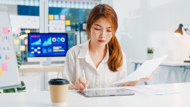 Succesvolle uitvoerende Aziatische jonge zakenvrouw slimme vrijetijdskleding tekenen, schrijven en gebruiken van pen met digitale tabletcomputer denken aan inspiratie zoeken ideeën werkproces in modern kantoor aan huis.