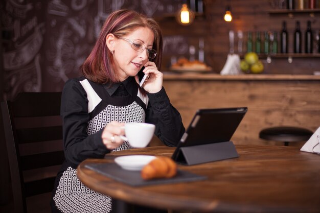 Succesvolle volwassen zakenvrouw die op haar telefoon praat terwijl ze een kopje koffie vasthoudt. Vintage restaurant