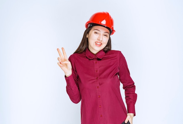 Succesvolle vrouwelijke architect in rode harde helm die staat en een overwinningsteken geeft.