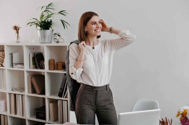 Succesvolle vrouwelijke beambte in grijze broek en licht shirt met glimlach vormt permanent op de werkplek.