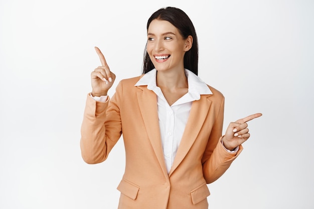 Succesvolle zakelijke vrouw in pak glimlachend wijzende vingers zijwaarts kijkend naar links naar logo bedrijfsadvertentie staande tegen een witte achtergrond