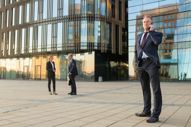 Succesvolle zakenman dragen kantoor pak, praten op een mobiele telefoon buitenshuis. Ondernemers en stad gebouw glazen gevel op achtergrond. Kopieer ruimte. Zakelijke communicatie concept