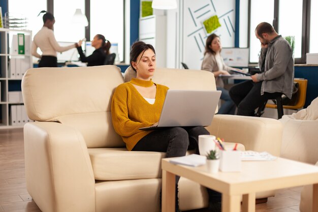 Succesvolle zakenvrouw die op laptop werkt aan opstartproject zittend op een gezellige bank