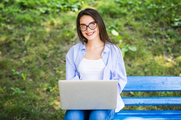 Sunny girl in blauw t-shirt zit op de bank in het park en gebruikt haar laptop