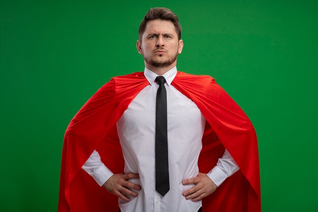 Super held zakenman in rode cape camera kijken met ernstige zelfverzekerde uitdrukking met armen op heup staande over groene achtergrond