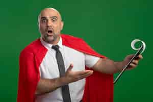 Gratis foto super held zakenman in rode cape met klembord met blanco pagina's wijzend met arm op klembord met verwarring uitdrukking staande over groene muur