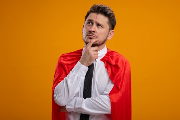 Super held zakenman in rode cape opzij kijken met peinzende uitdrukking denken staande over oranje achtergrond