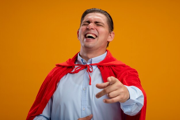 Super held zakenman in rode cape wijzend met wijsvinger op camera lachen staande over oranje achtergrond