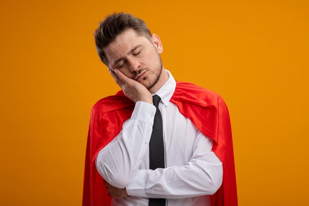 Super held zakenman in rood capeleaning hoofd op zijn handpalm op zoek moe en verveeld staande over oranje muur