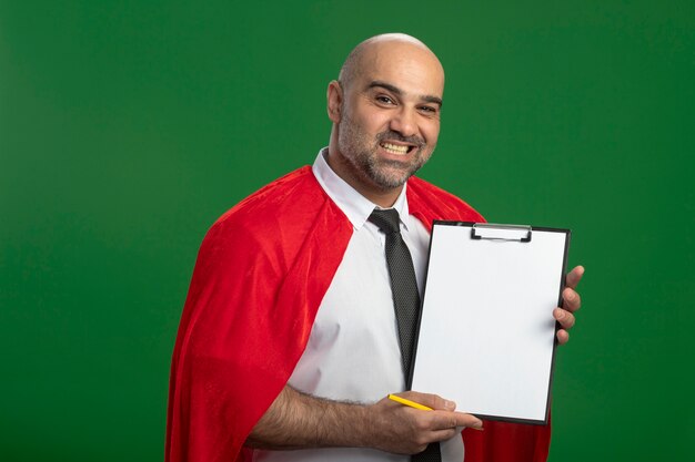 Super heldzakenman in rode cape die klembord met lege pagina's toont die voorzijde met glimlach op gezicht bekijken die zich over groene muur bevinden