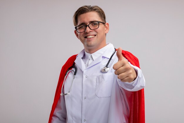 Superheld dokter man met witte jas in rode cape en bril met stethoscoop om nek met glimlach op gezicht duimen opdagen staande over witte muur