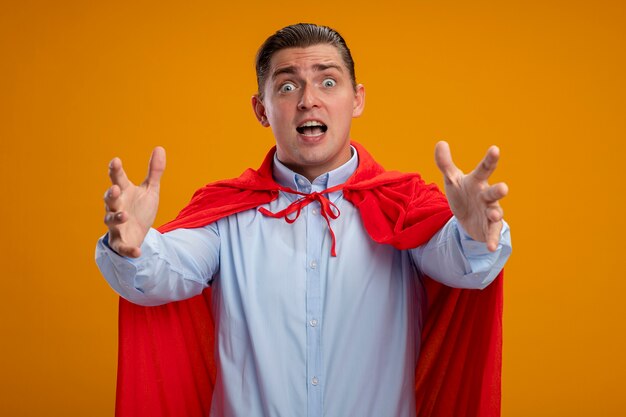 Superheld zakenman in rode cape met gekke blik van verrassing, gek, verbaasd en verrast met armen uitgestrekt over oranje muur