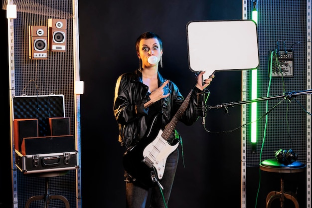 Gratis foto superstervrouw wijzend op een leeg reclamebord terwijl ze op een elektrische gitaar speelt en zich voorbereidt op een rockconcert. rebellenmuzikant met grunge-stijl die heavy metal-liedjes uitvoert in de studio