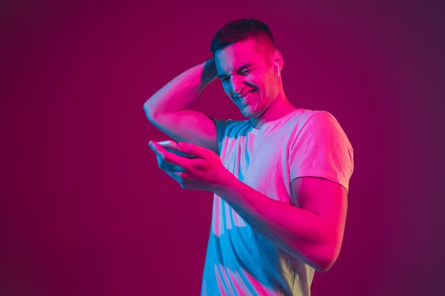 Surfen, wedden, kijken, selfie. Portret van een blanke man geïsoleerd op roze-paarse muur in neonlicht. Mannelijk model met apparaten. Concept van menselijke emoties, gezichtsuitdrukking,