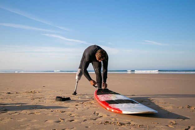 Surfer in wetsuit en kunstledemaat, boord aan zijn enkel op zand bindend