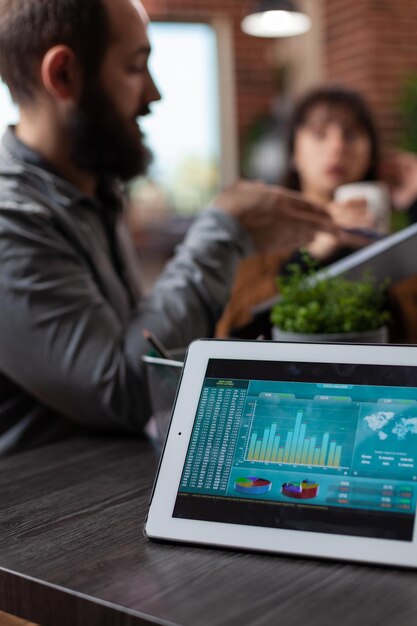 Tabletcomputer met bedrijfsstatistieken op het scherm dat op tafel staat in het opstartkantoor. Millennial multi-etnische zakenmensen bespreken marketingstrategie die werken aan zakelijke samenwerking