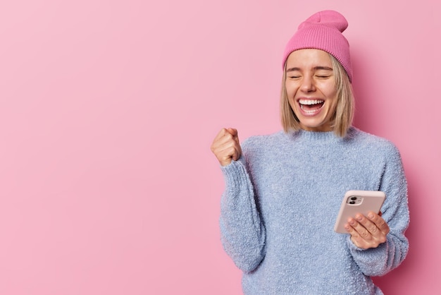 Taille omhoog geschoten van gelukkige positieve blonde vrouw balt vuist van vreugde verheugt zich goed nieuws maakt gebruik van mobiele telefoon voor communicatie gekleed in casual kleding geïsoleerd over roze achtergrond lege ruimte weg