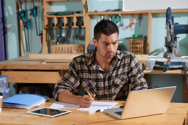 Taille omhoog geschoten van het knappe timmerman planningswerk aangaande volgende project bekijkend laptop met in hand potlood