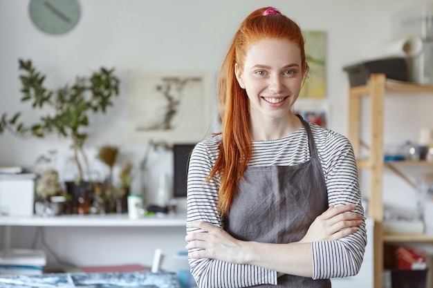 Gratis foto taille omhoog geschoten van vrolijke jonge kaukasische vrouwelijke creatieve kunstenaar, ontwerper of kunstenaar met lang rood haar en sproeten die uitdrukking op haar gezicht hebben geïnspireerd tijdens het werken in haar moderne werkplaats