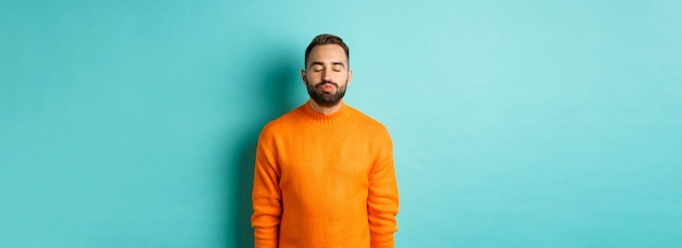 Gratis foto taille omhoog schot van jonge man tuit lippen en gesloten ogen wachtend op kus staande in oranje trui ag