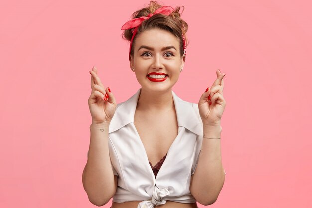 Taille portret van pinup mooi vrouwelijk model met rood geschilderde lippen en manicure, retro-stijl heeft, vingers kruist met positieve expressie, hoopt op iets, heeft doel in het leven