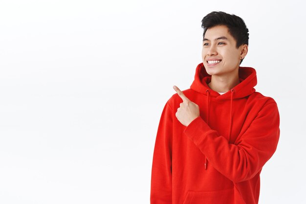 Taille-up portret van aantrekkelijke gelukkige hipster man in rode hoodie, oorbel, tevreden glimlachend en wijzend op zoek naar de linker bovenhoek met opgetogen