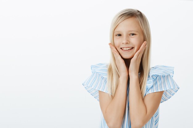 Taille-up portret van positief tevreden schattig vrouwelijk kind met blond haar, breed glimlachend van ontvangen compliment en handpalmen op de wangen vasthoudend, zich geweldig en schattig voelen