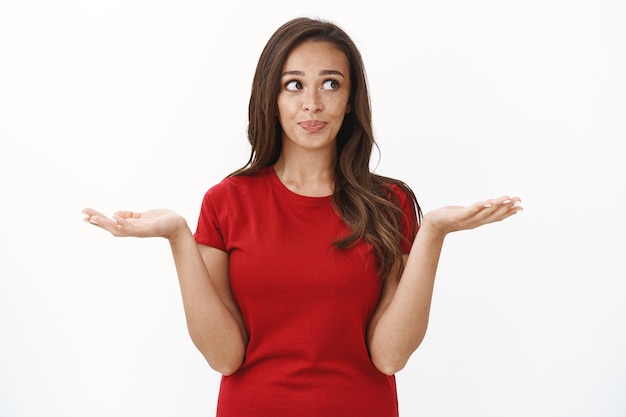 Taille-up shot schattige domme brunette vrouw in rood casual t-shirt, schouderophalend handen omhoog ondervraagd, grijns en wegkijken keuze maken, nadenken tussen varianten, staande witte muur