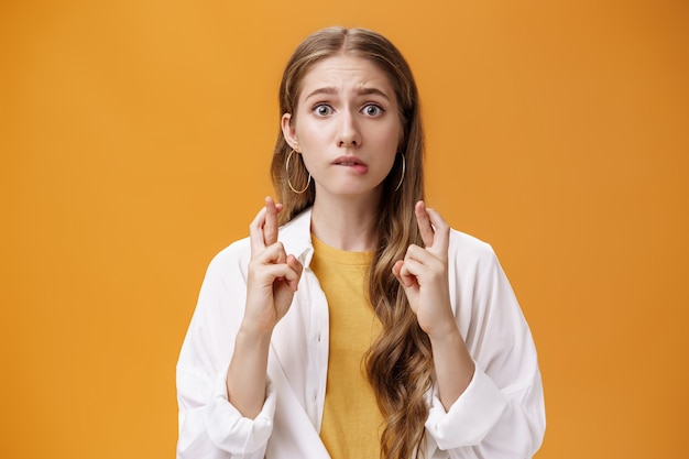 Taille-up shot van bezorgd en opgewonden schattige jonge vrouw in blouse over t-shirt met golvend lang haar lip bijten nerveus hoopvol kijkend naar camera die vingers kruist voor geluk tijdens het bekijken van resultaten.