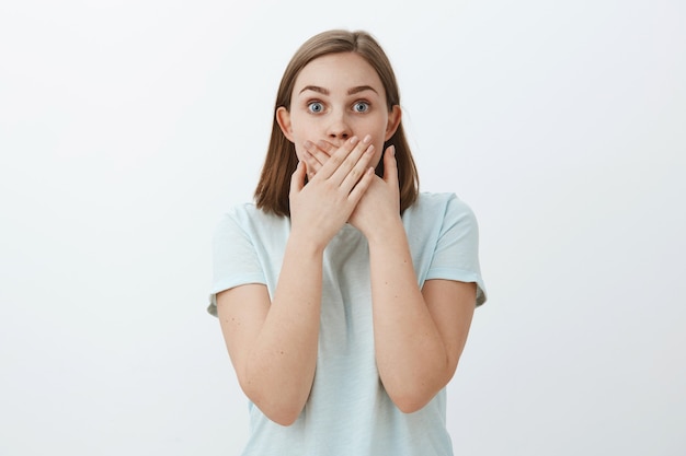 Taille-up shot van een verbaasde verbijsterde vrouw die een schokkend gerucht leert dat de mond bedekt met beide handpalmen starend verbaasd onder de indruk en opgewonden poseren voor de grijze muur