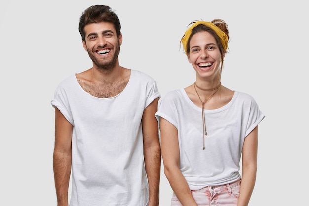 Gratis foto taille-up shot van gelukkige vrouwelijke en mannelijke kerels gekleed in een wit mockup-t-shirt, breed glimlachen, in hoge geest zijn, dicht bij elkaar staan, geïsoleerd over de muur
