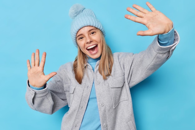 Taille-up shot van gelukkige zorgeloze vrouw houdt handpalmen omhoog naar camera kantelt hoofd roept graag warme wintermuts grijze jas heeft plezier geïsoleerd op blauwe achtergrond ziet er vol vreugde uit