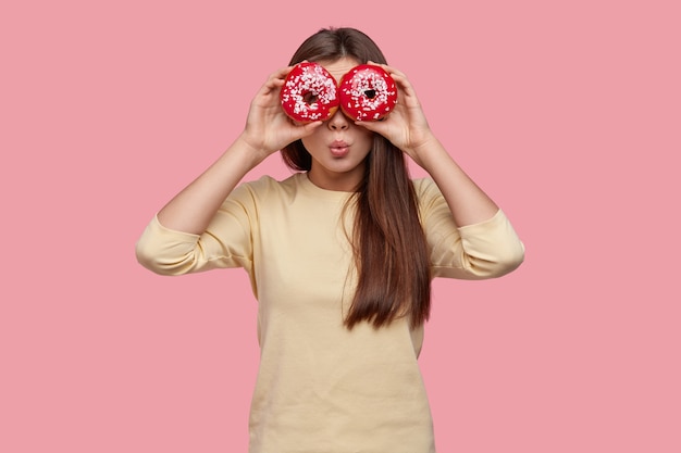 Taille-up shot van mooie jonge dame heeft betrekking op ogen met twee rode donuts, draagt vrijetijdskleding, staat op roze achtergrond